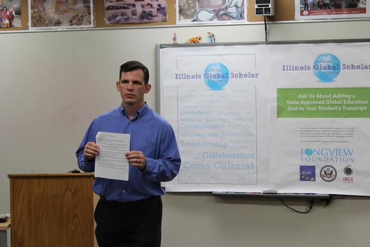 Seth Brady, social studies teacher, stands in his classroom with posters advertising the Global Scholar Program behind him.